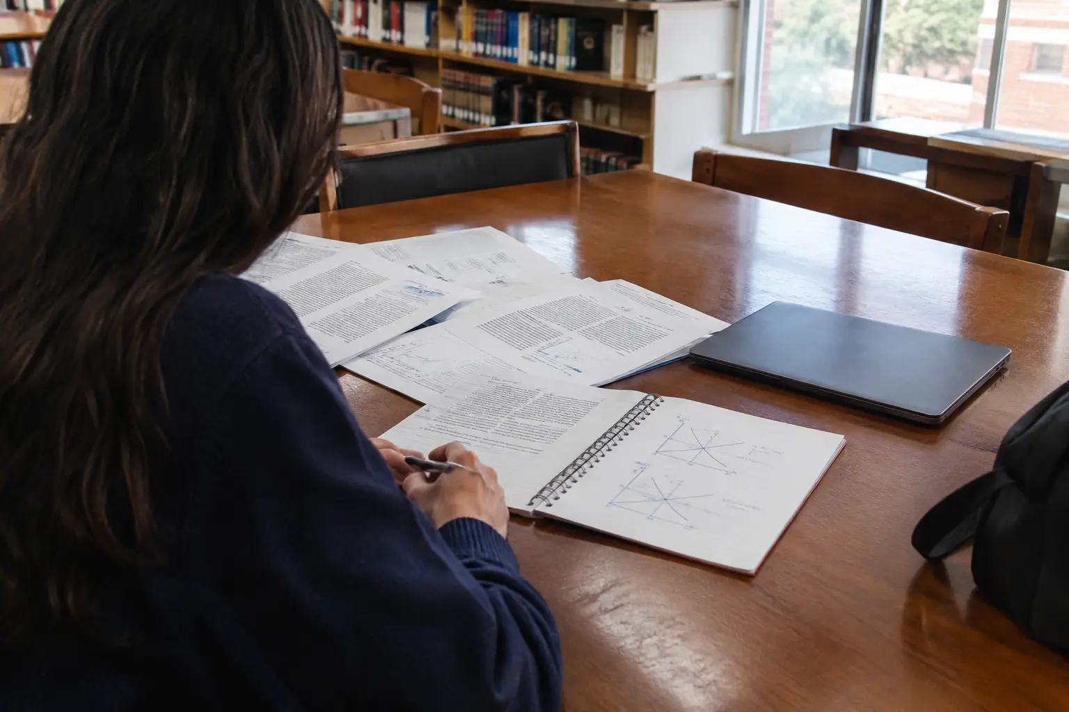 Estudiante de economía revisando artículos académicos impresos y notas con curvas de oferta y demanda